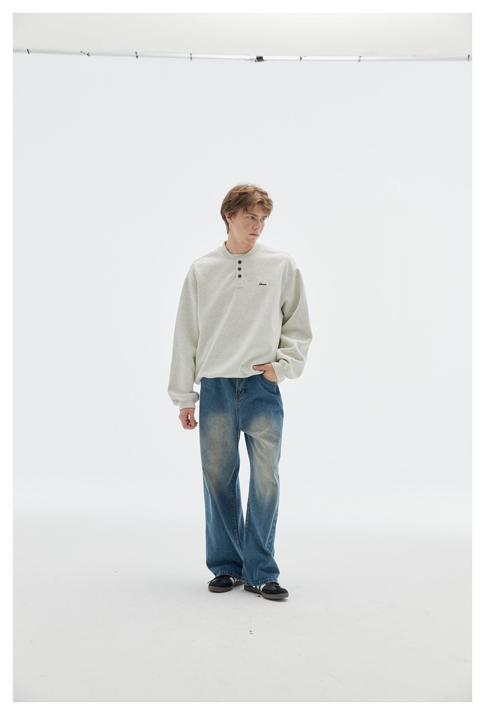 Model wearing an off-white henley sweatshirt with loose blue jeans, shown full length against a light studio background.