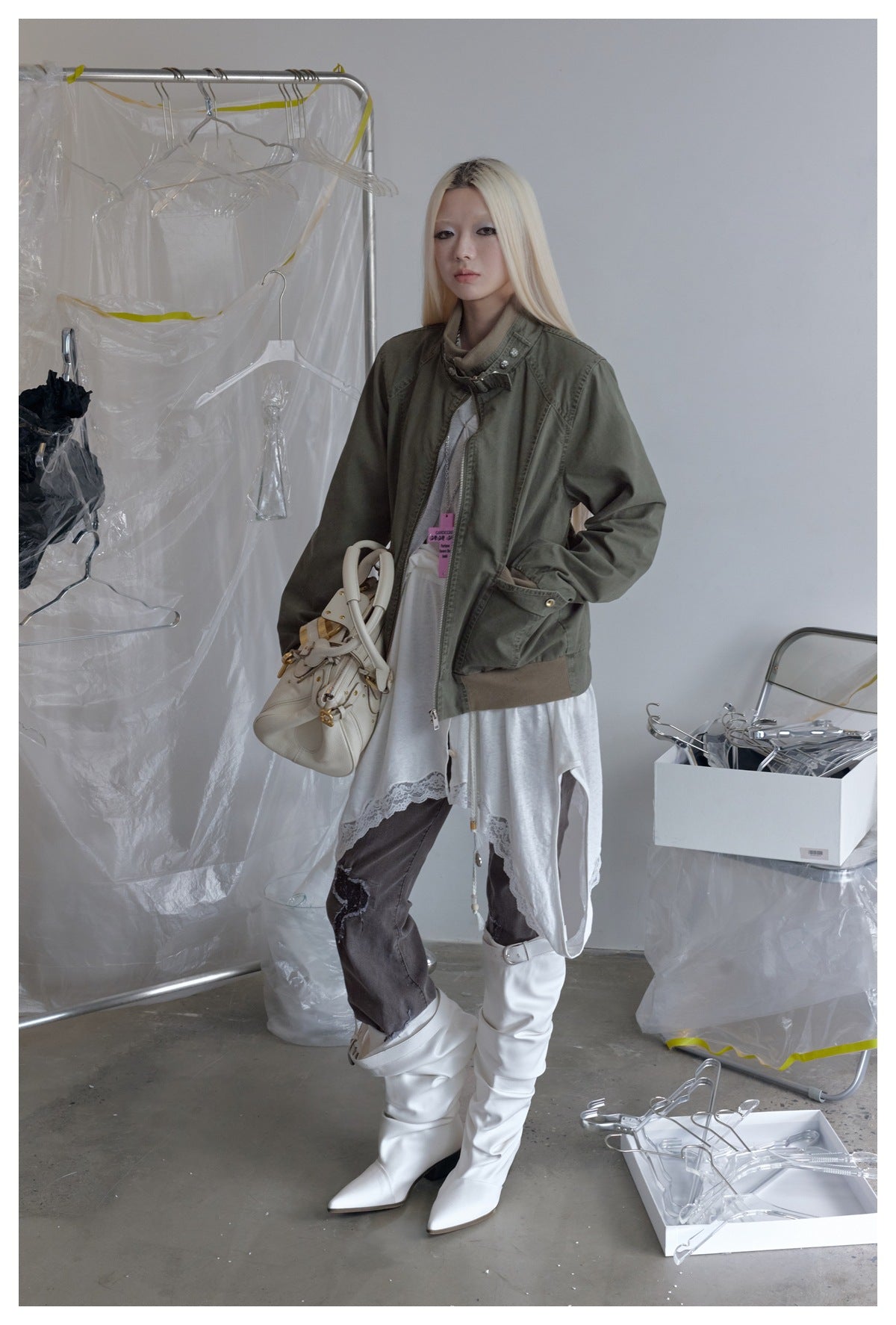 Model in the olive green zip jacket with tan ribbed hem, styled with a white lace-trim top, white boots and a handbag