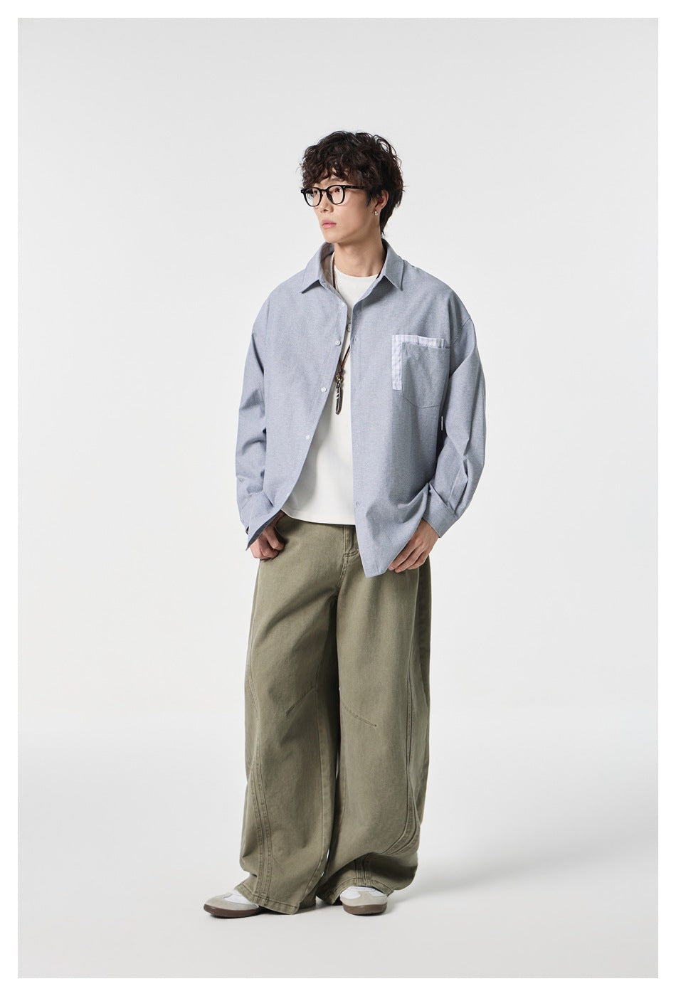Full-length model wearing a loose gray-blue button-up shirt with a plaid-trimmed chest pocket.