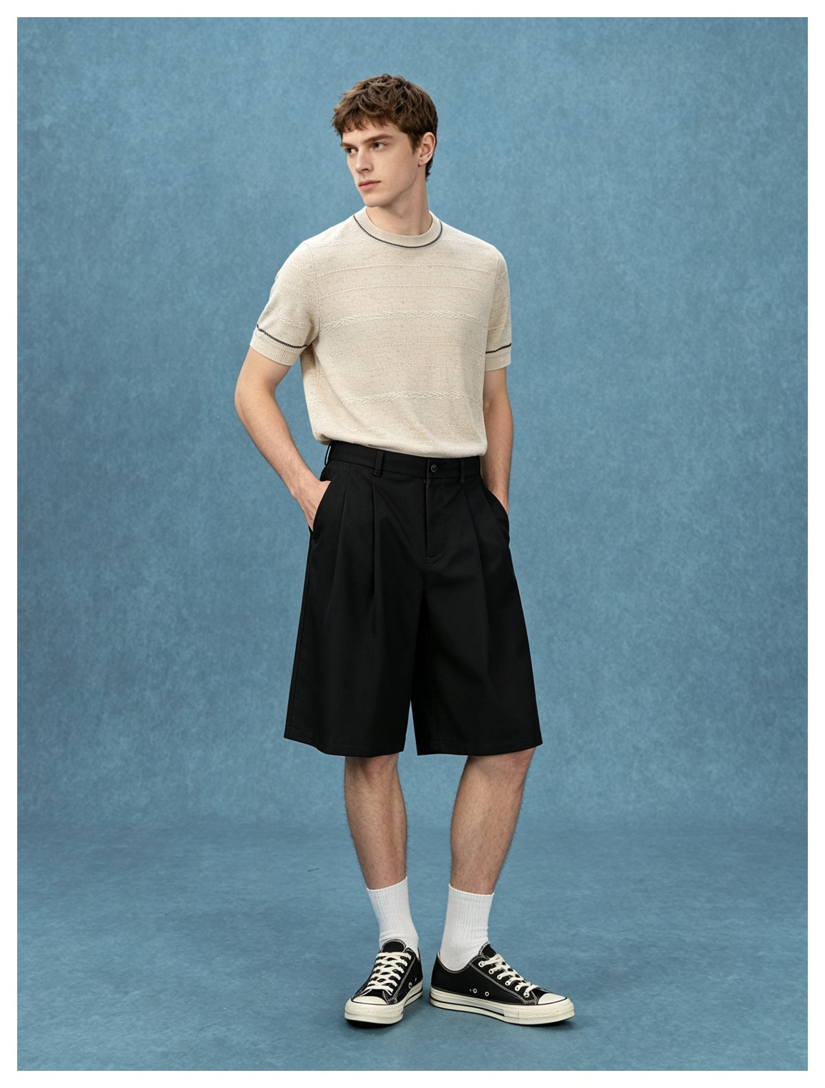 Man wearing khaki short-sleeve knit top with contrast stitching, black pleated shorts and sneakers against blue backdrop