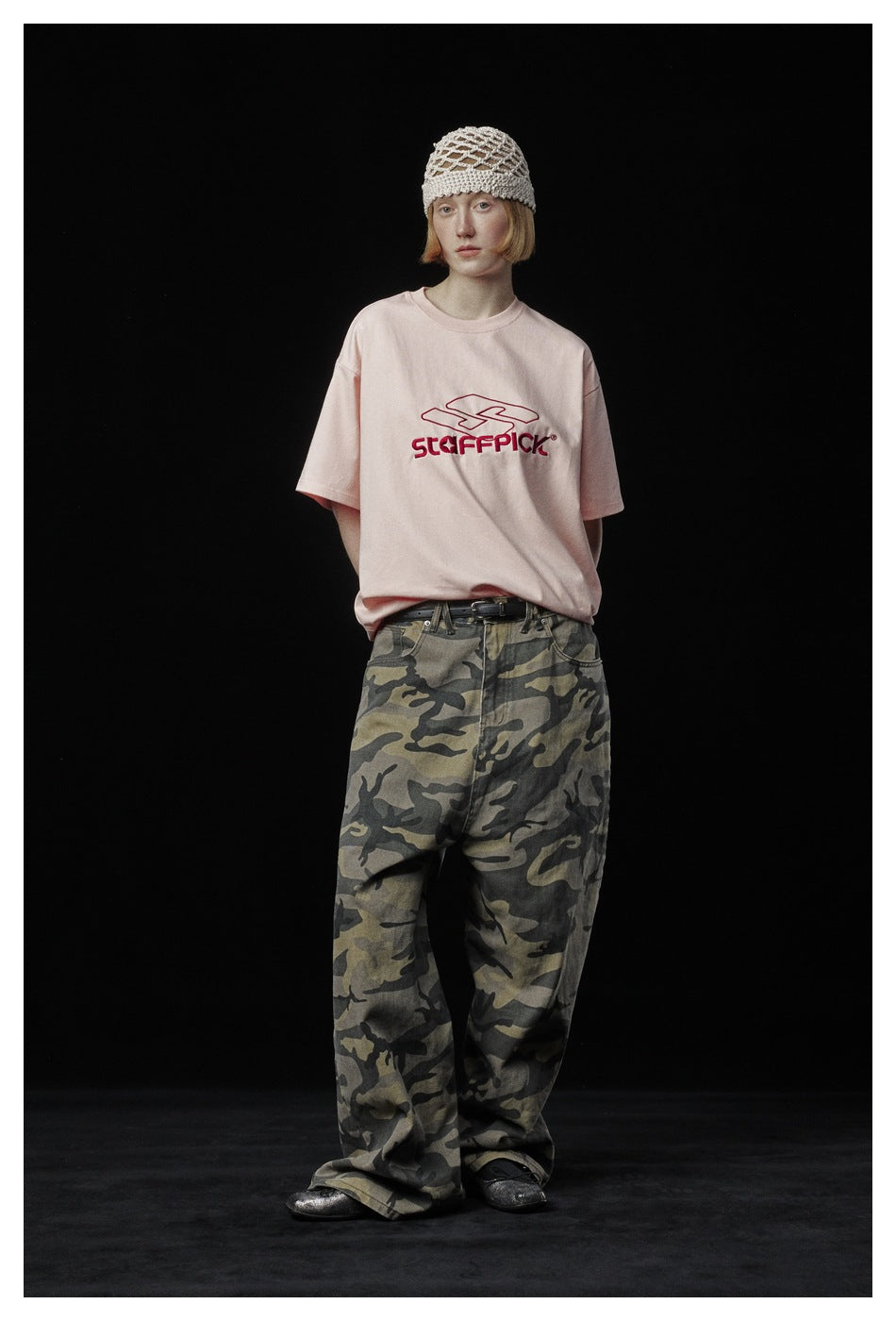 Model wearing a pale pink graphic T-shirt, loose camouflage trousers, and a cream open-knit hat.