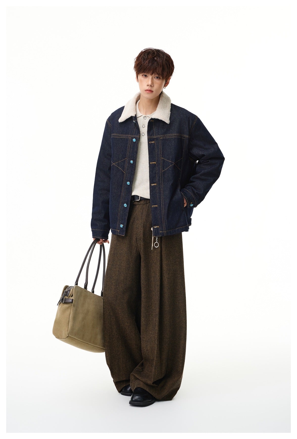 Model wearing denim jacket with cream fleece-like collar over a light polo, brown wide-leg trousers, holding a tan bag