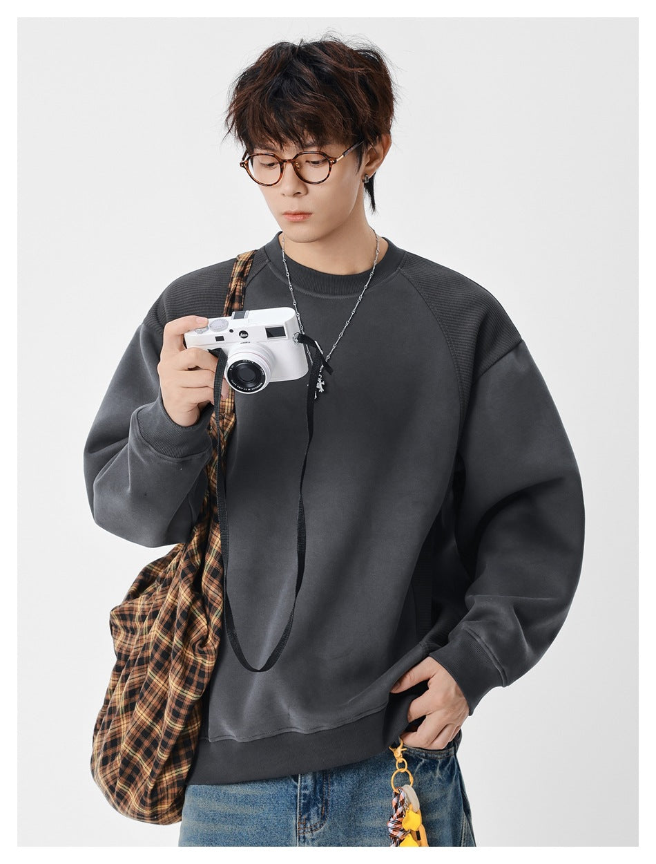 Man wearing gray crewneck sweatshirt with ribbed raglan panels, holding a camera, with a plaid bag and jeans