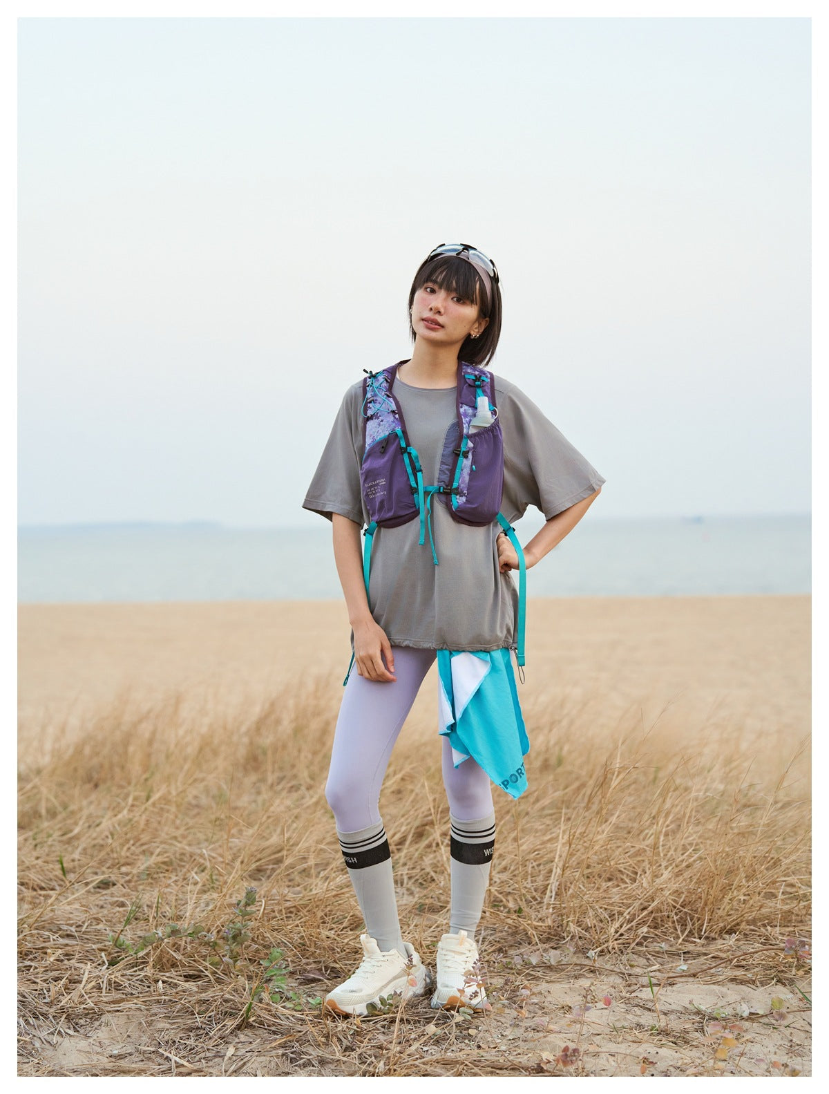 Woman wearing the purple camo mesh vest with teal straps over a gray tee and lilac leggings on a beach