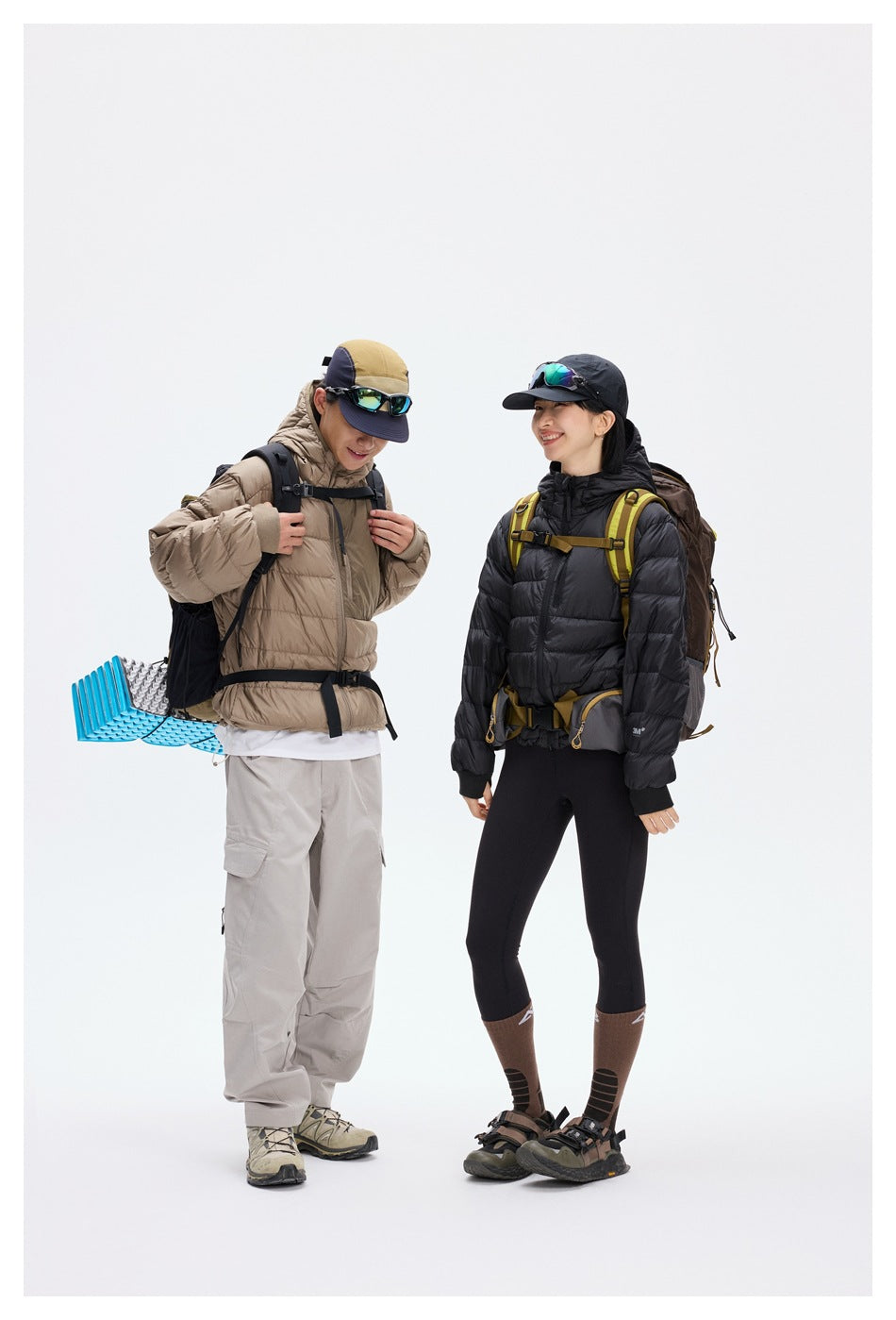 Two hikers wear taupe and black hooded puffer jackets with backpacks, shown full length against a white studio background.