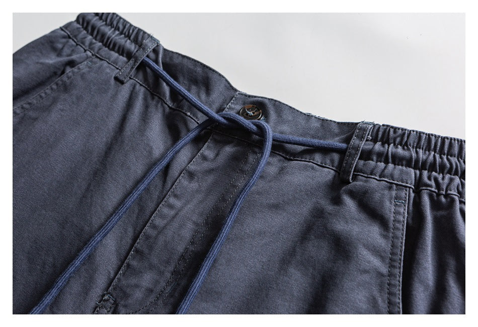 Close-up of slate blue trousers showing the elastic waistband, button closure, belt loops, and drawstring.