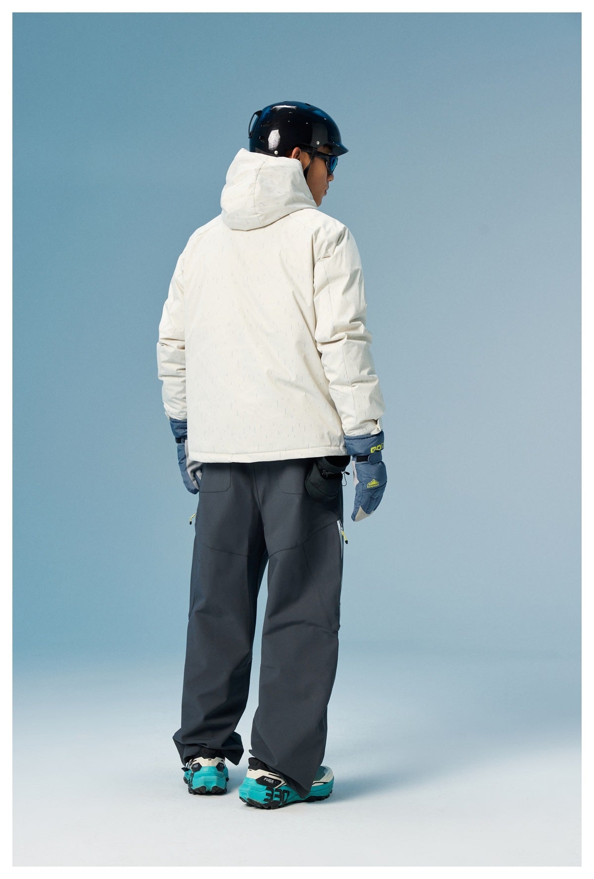 Rear three-quarter view of a model wearing a white hooded ski jacket and dark snow pants with blue gloves.