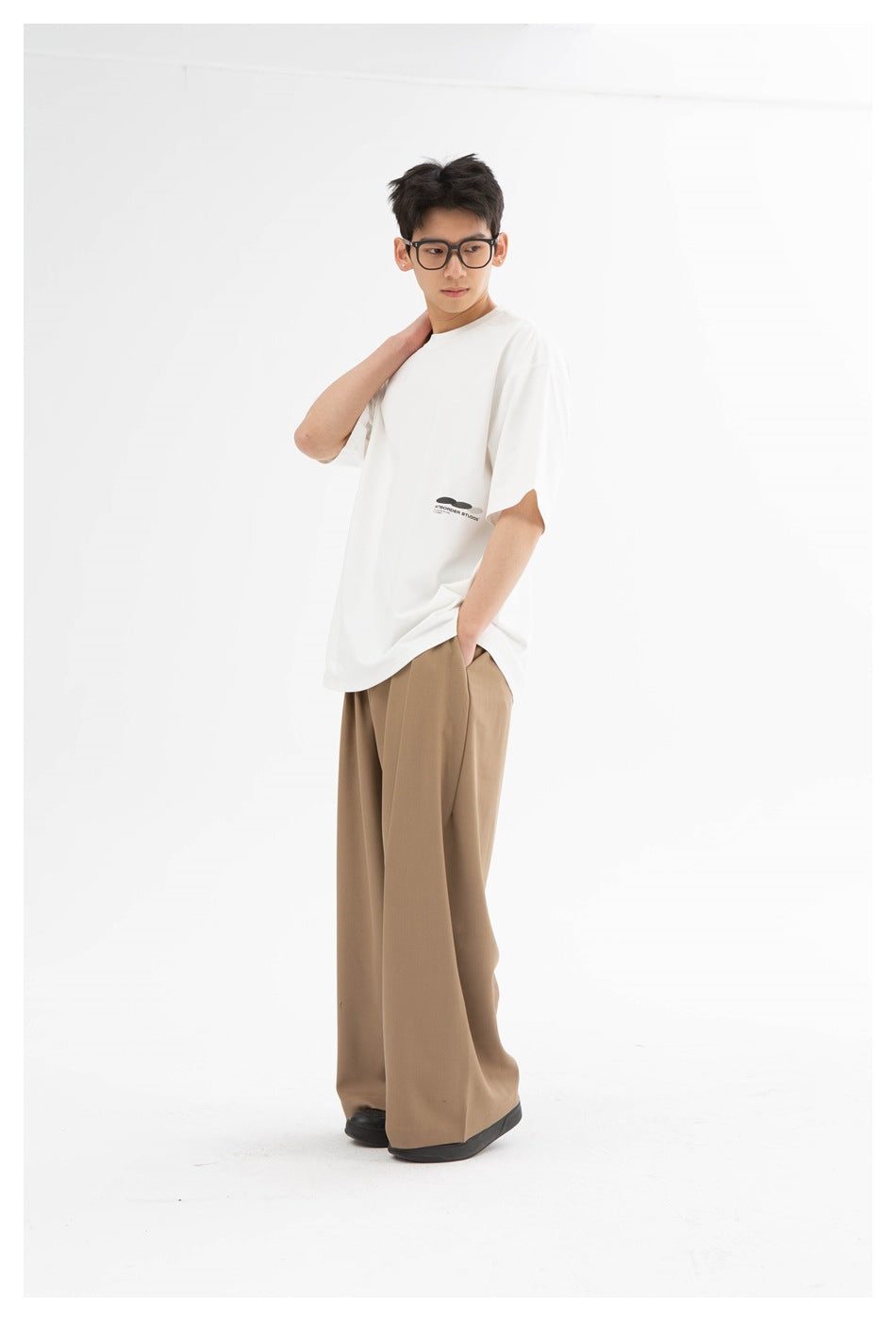 Model in a white oversized t-shirt with small lower-hem print, wide-leg tan trousers and black shoes, hand at neck