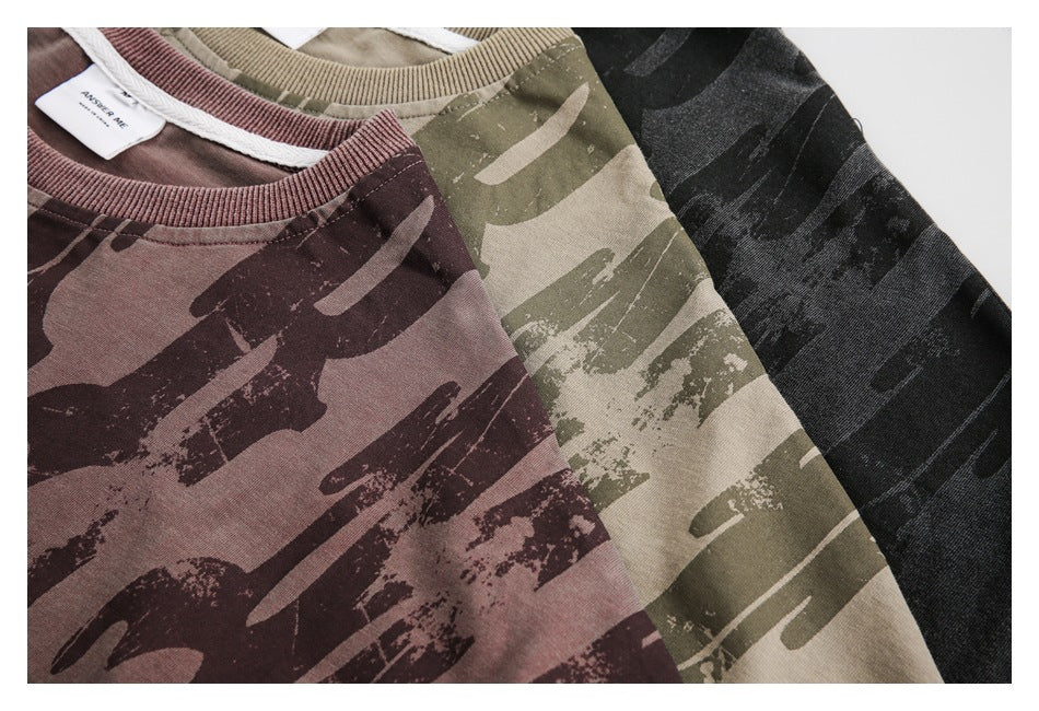 Close-up of mauve, khaki, and black T-shirts with abstract brushstroke camouflage prints and ribbed collars.