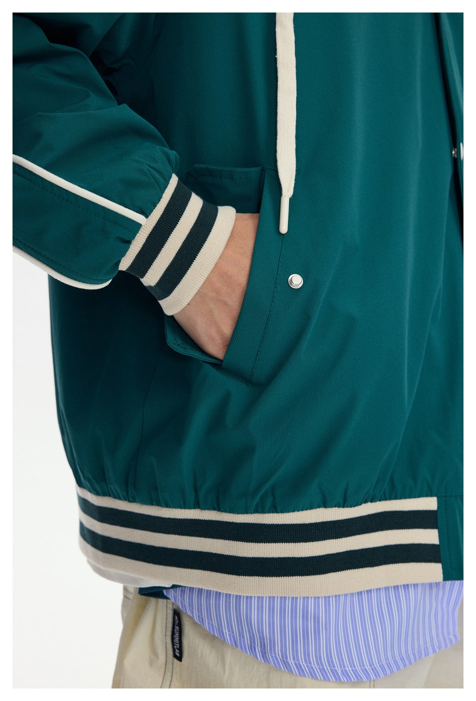Close-up of a teal jacket showing its snap pocket, cream drawcord, striped cuffs and hem, and striped shirt underneath.
