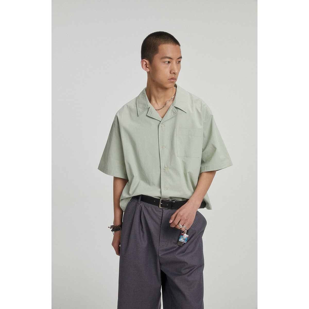 Model wearing a sage-green short-sleeve button-up shirt with a pointed collar and chest pocket.