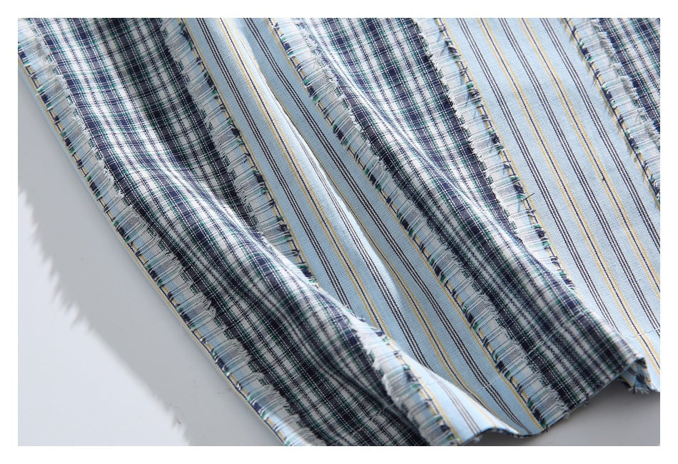Close-up of light blue fabric with alternating dark plaid and narrow striped bands, with fringed edges.