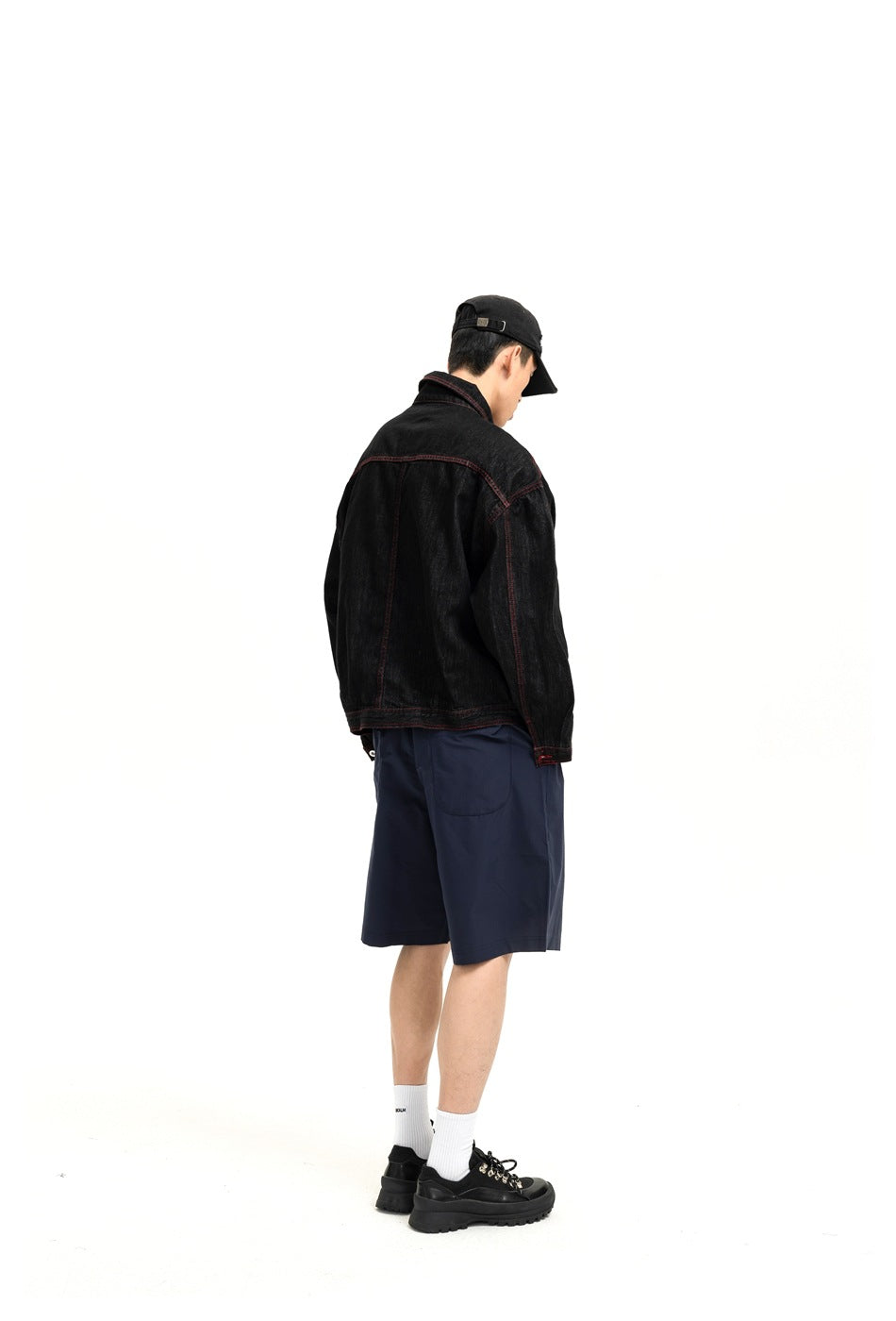 Rear view of a model wearing a black denim jacket with red stitching and navy shorts.