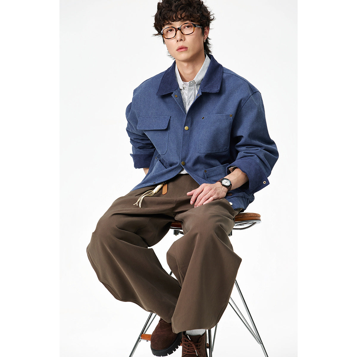 Seated model wearing a blue button-front work jacket with four pockets and wide brown trousers.