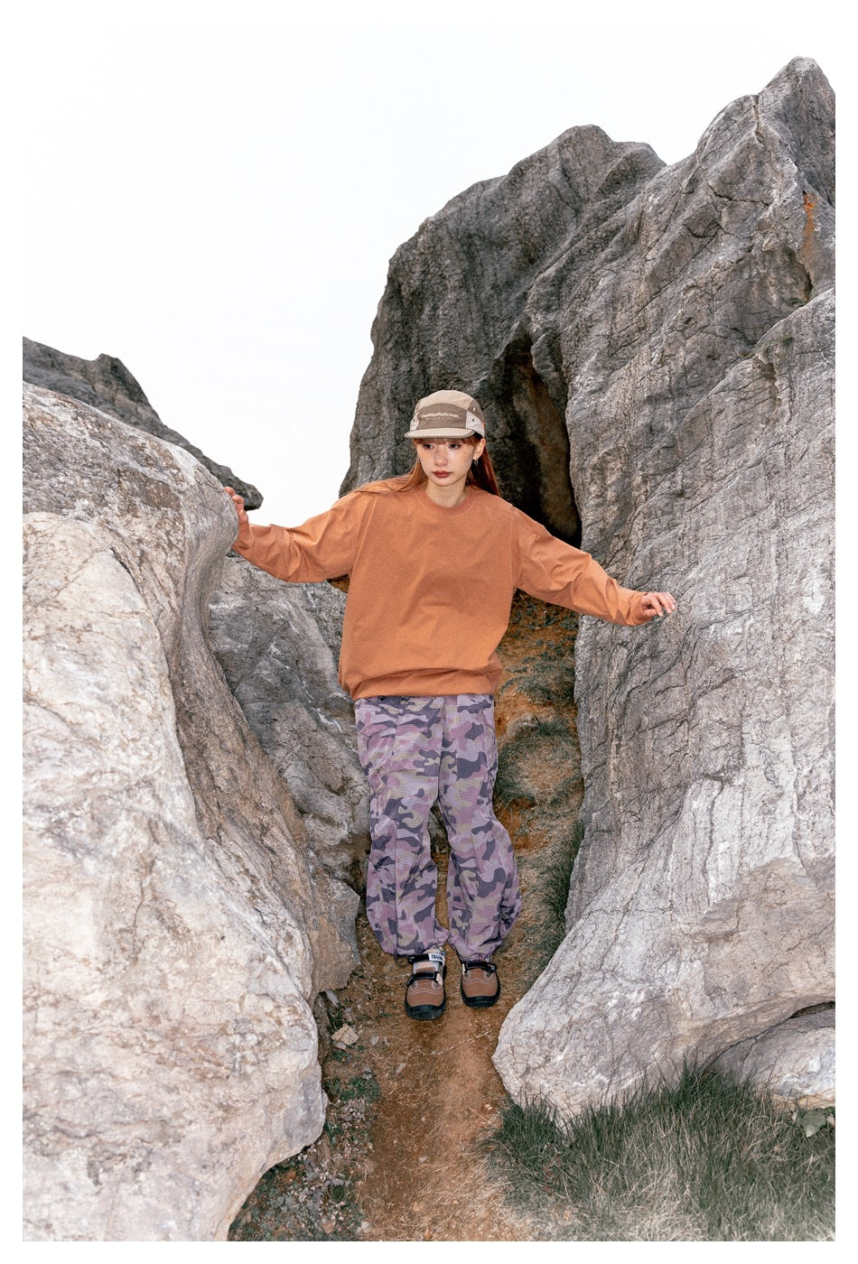 Model wearing a rust-orange sweatshirt and purple camouflage pants, standing with arms out among rocks.