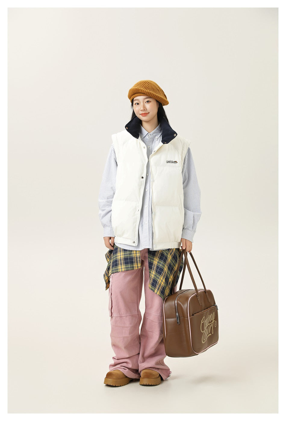 A model wears a white padded vest with a navy collar over a striped shirt, with pink trousers and a mustard knit cap.