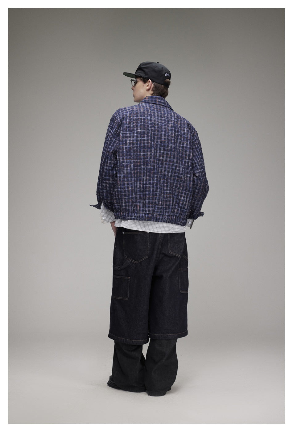Back view of a model wearing a dark navy plaid jacket with an elastic hem and black denim shorts over dark pants.