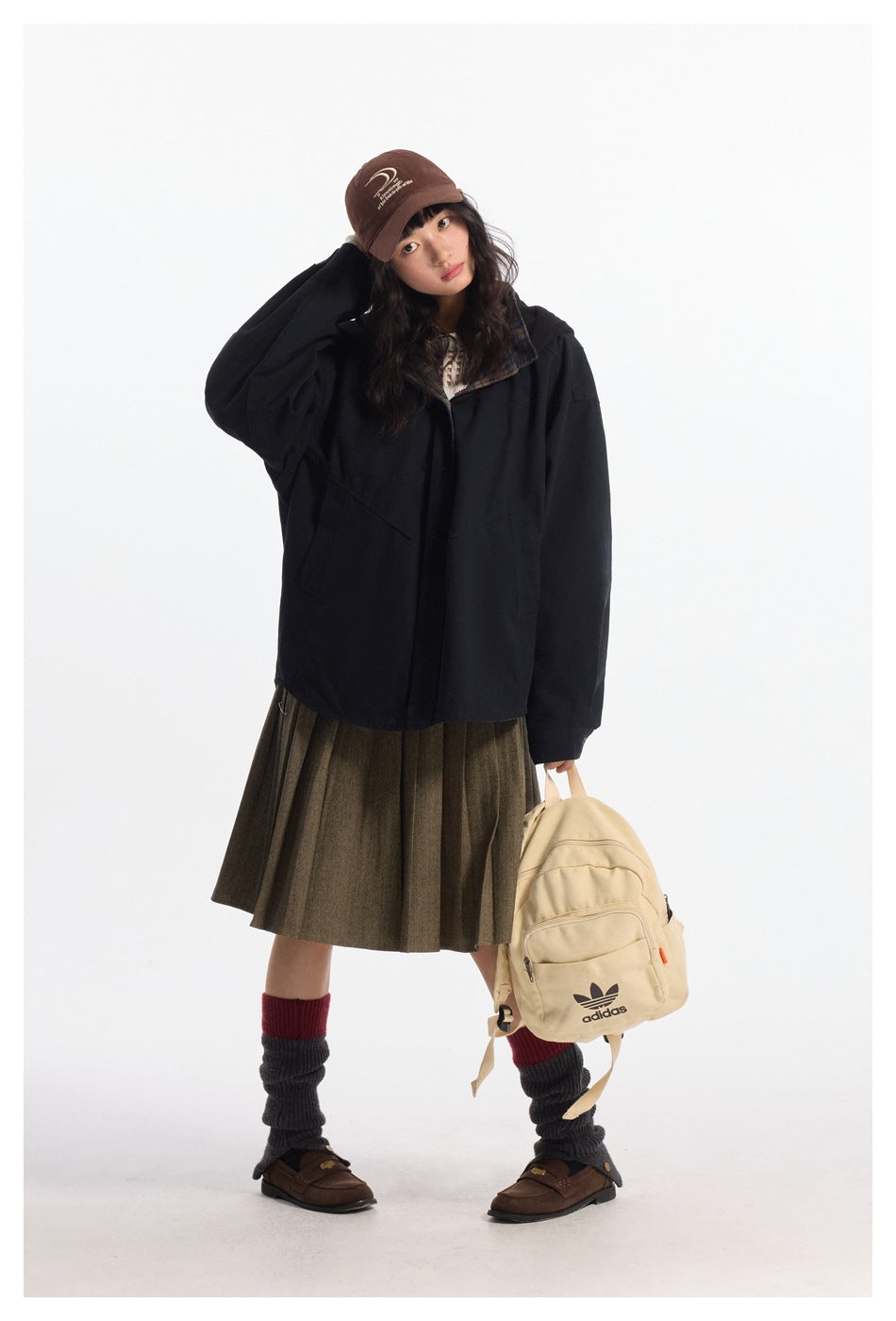 Full-length model wearing an oversized black jacket with a plaid collar, brown skirt, and carrying a cream backpack.