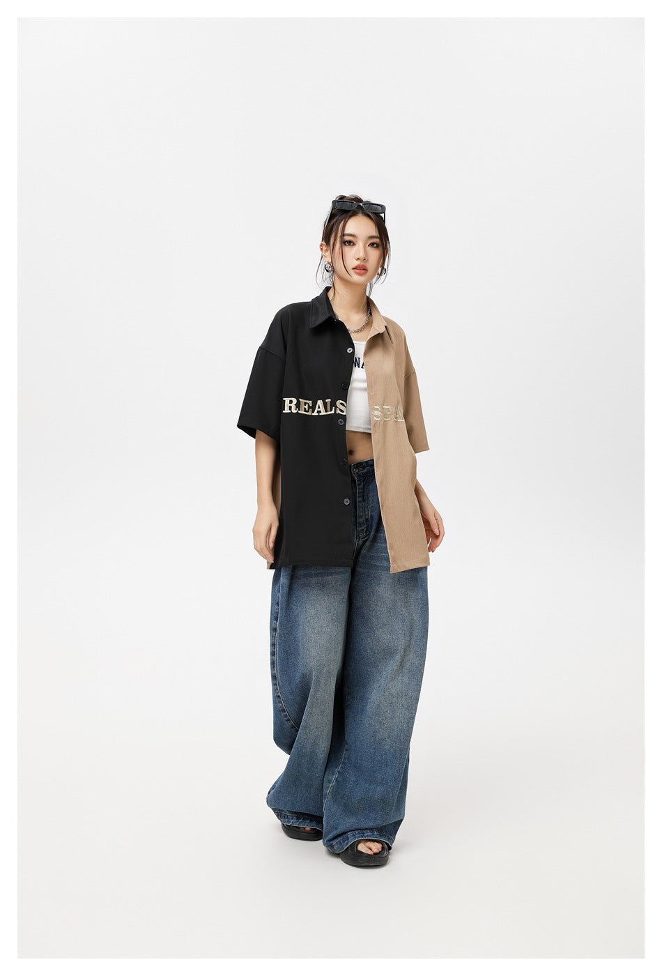 Model wearing a black-and-tan embroidered button-up shirt over a white crop top with loose blue jeans.