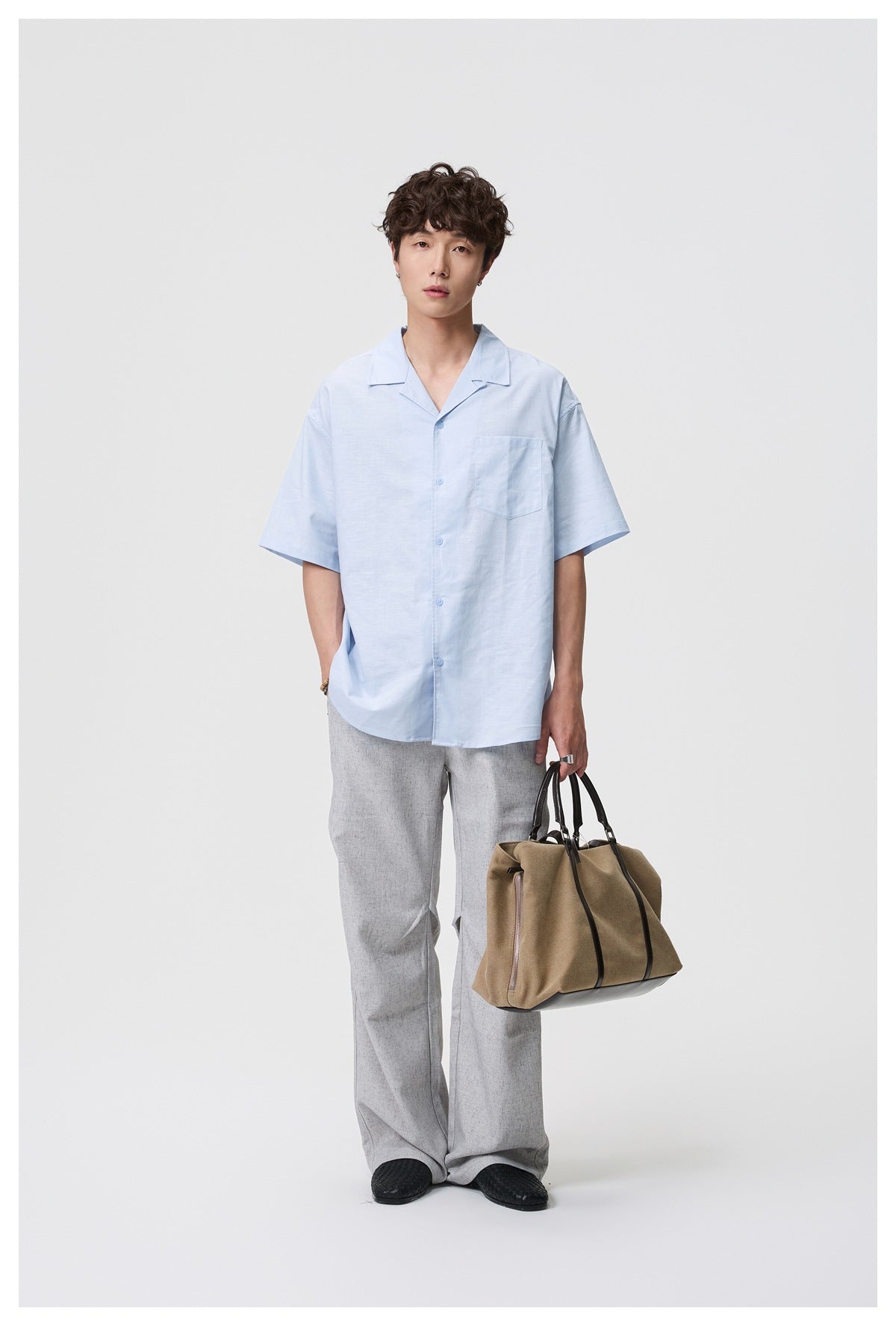 Man wearing a light blue short-sleeve camp collar shirt with light gray pants, holding a tan tote bag