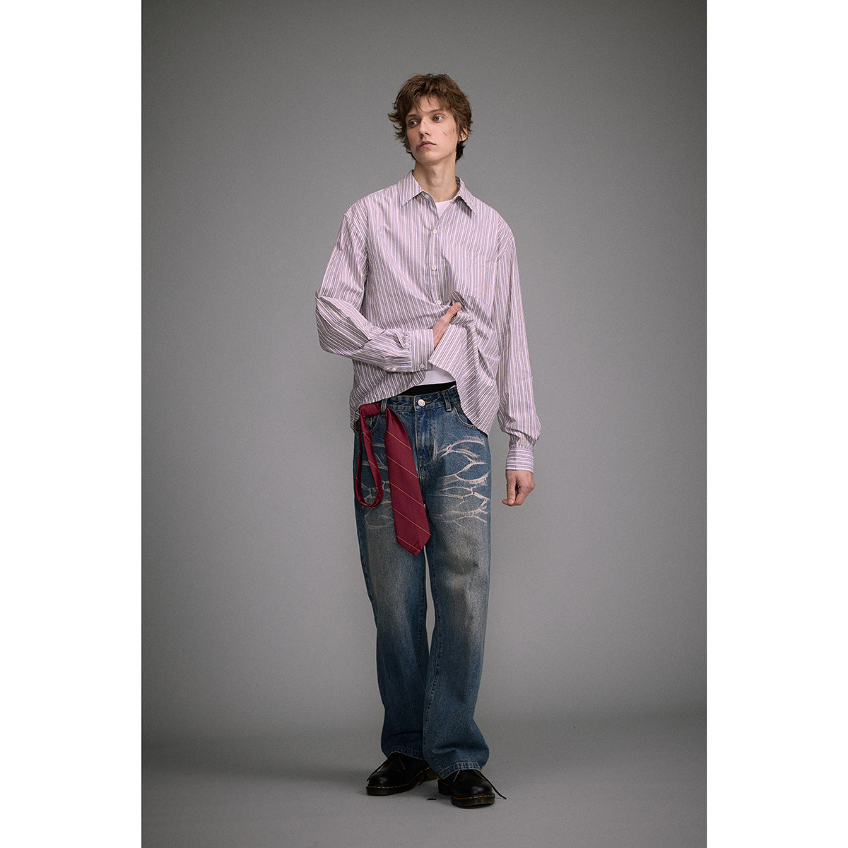 Model wearing a loose pink-and-white striped button-up shirt with a chest pocket, styled with jeans and a red tie.