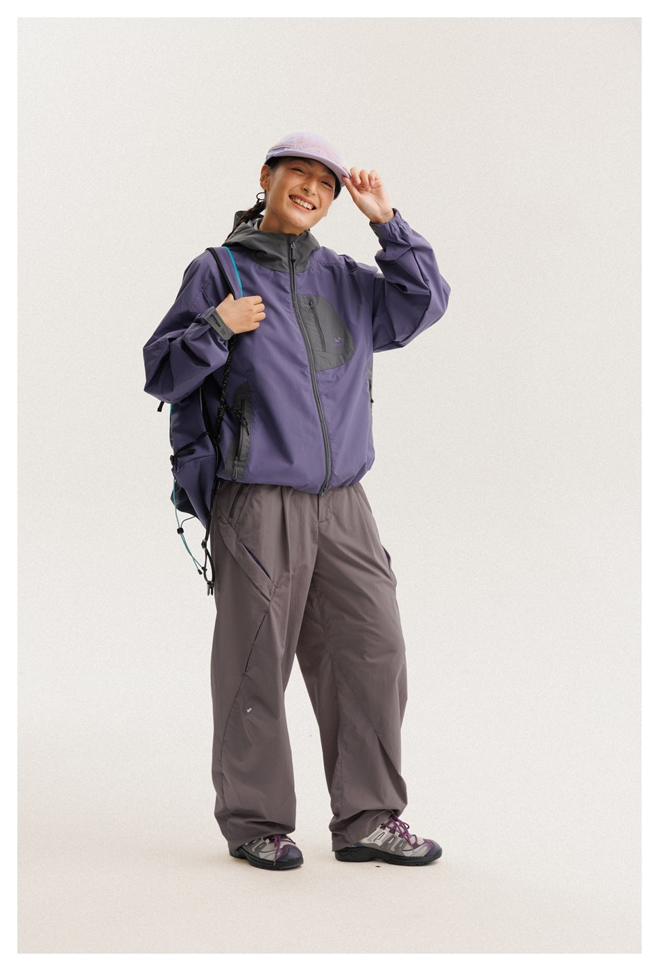 Model wearing a purple and charcoal hooded jacket with loose gray pants and a lavender cap, carrying a backpack.