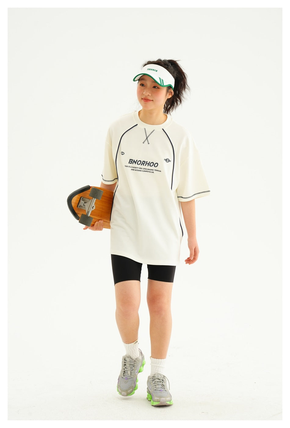 Model walking in an ivory oversized graphic T-shirt with dark piping and black shorts, carrying a skateboard.