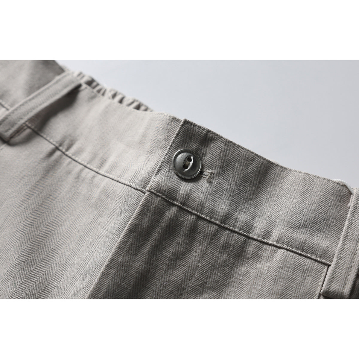 Close-up of gray herringbone trousers showing a dark button, waistband stitching, and partial inner-waist lettering.