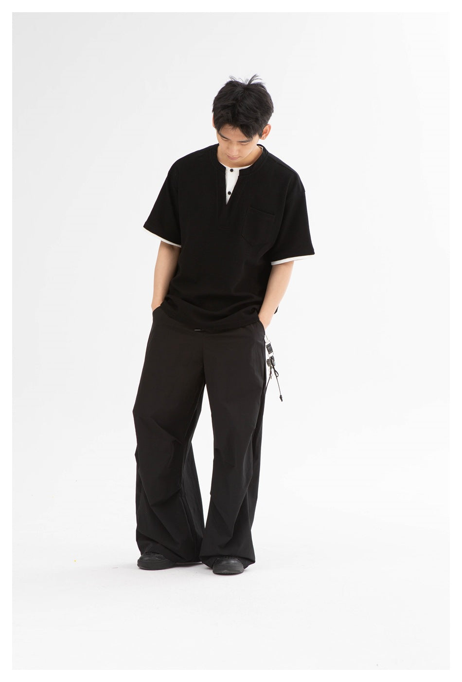 Man in a black short-sleeve henley with white trim and loose black trousers, shown looking down.