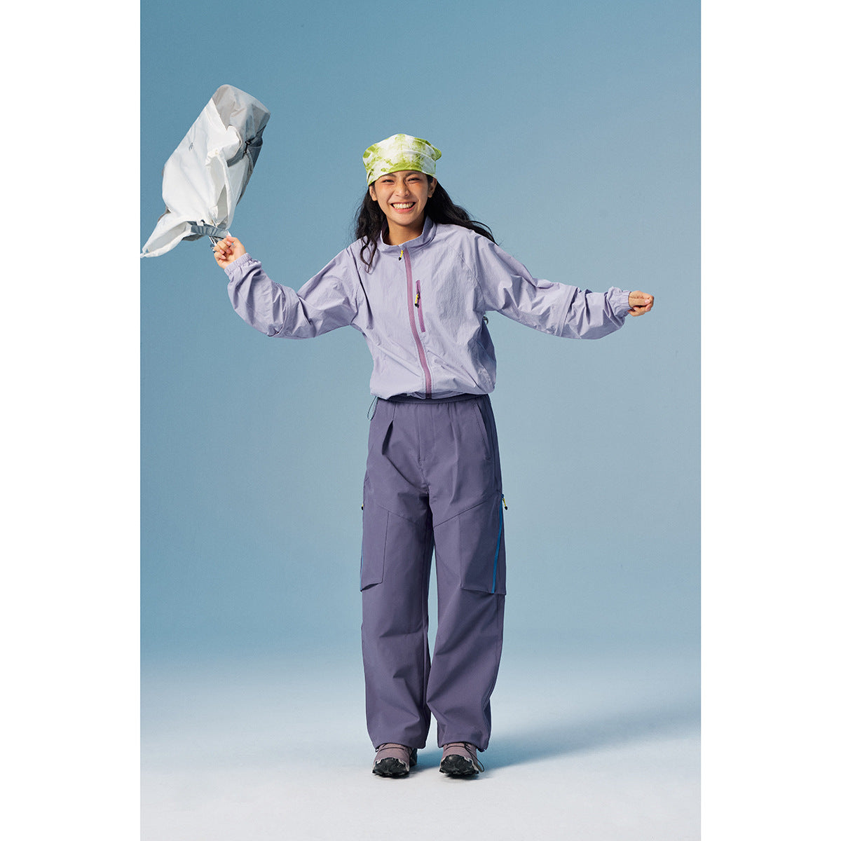 Woman in a lavender zip jacket and matching loose pants, holding a light-colored bag against a blue backdrop.
