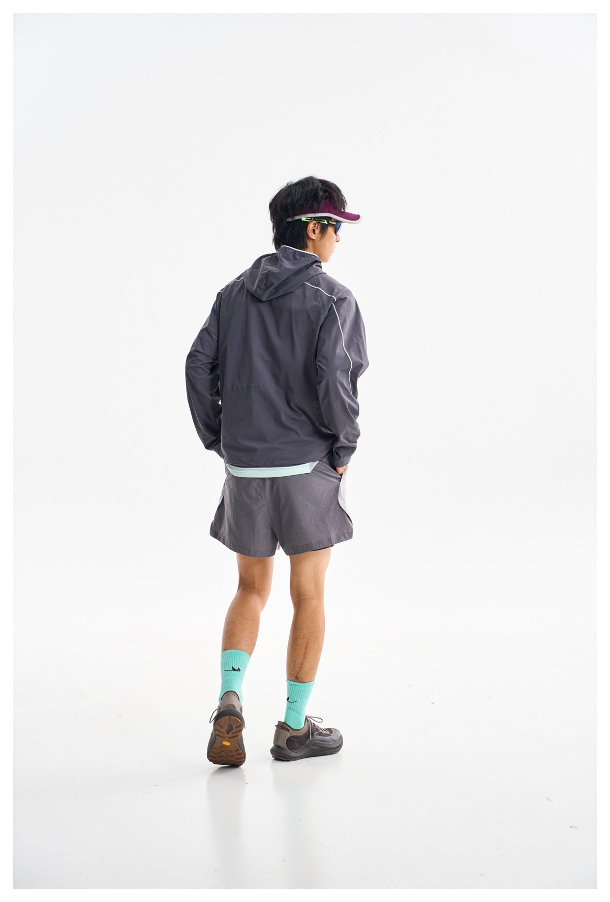 Rear view of a model wearing a charcoal hooded jacket with pale piping and matching dark shorts.