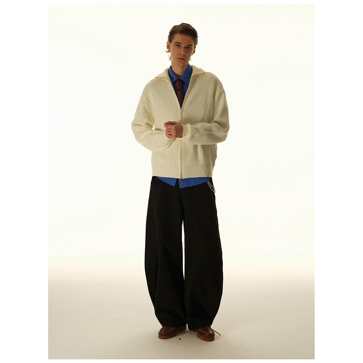 Model wearing a cream ribbed zip-up cardigan over a blue shirt and tie with wide black trousers.