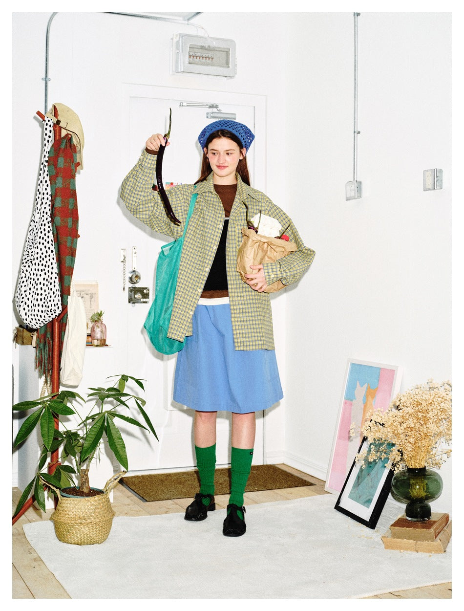 Model wearing a yellow-and-blue plaid shirt and blue skirt, holding a paper bag and a long dark vegetable.