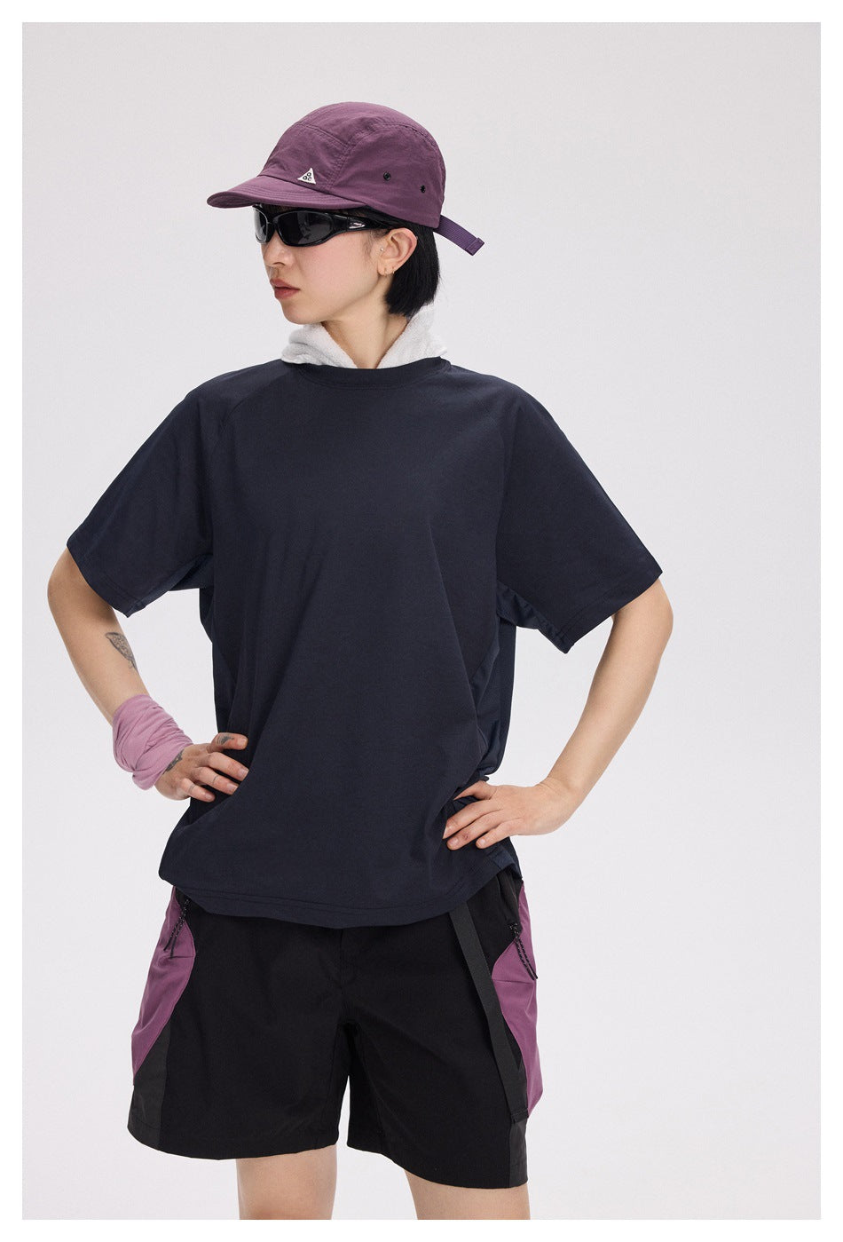 Model wearing a loose dark navy short-sleeve T-shirt and black shorts with purple side panels, hands on hips.