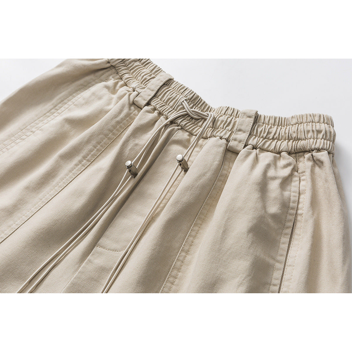 Close-up of beige shorts showing the gathered elastic waistband, drawstring toggles, and stitched panels.