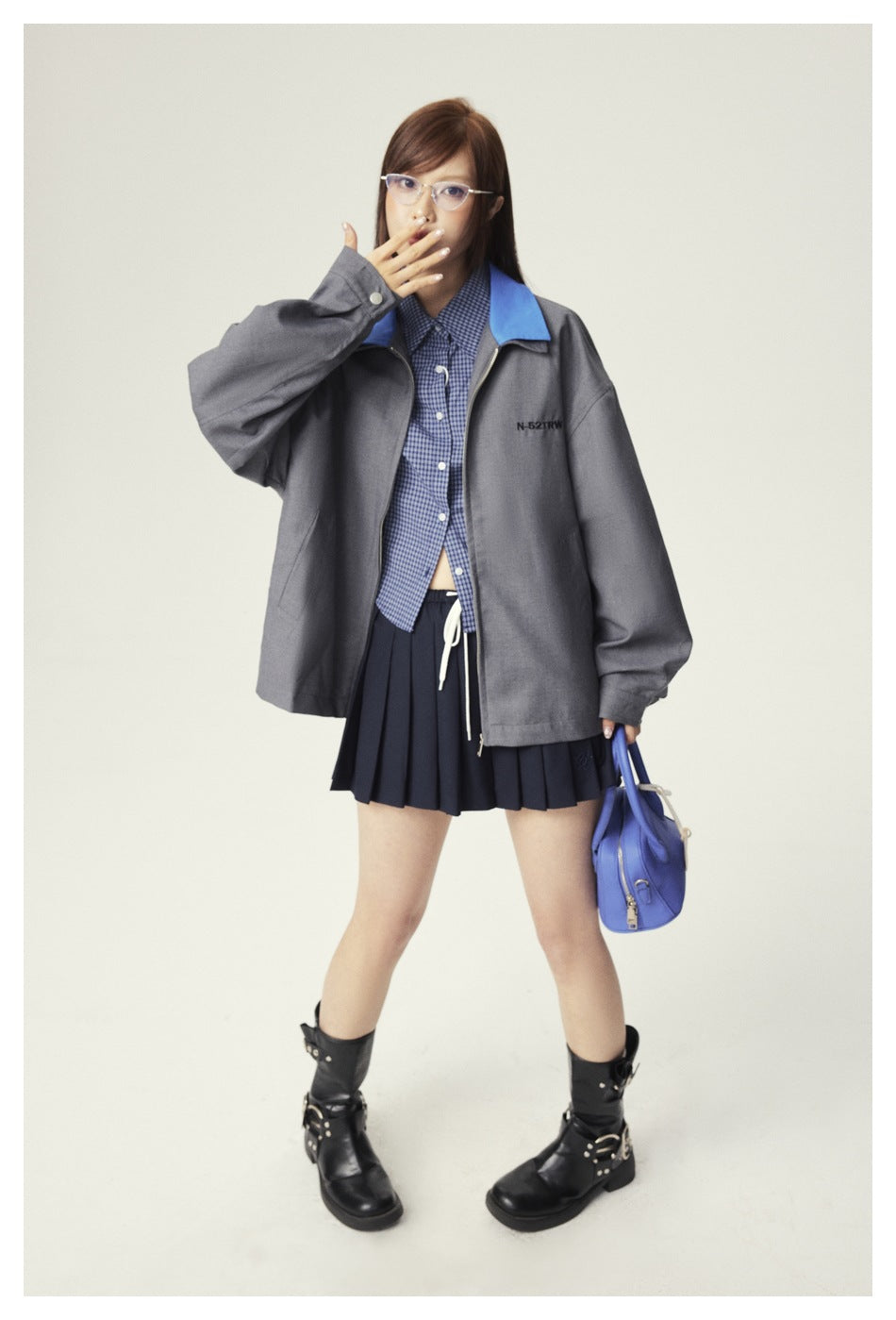 Model wearing a gray zip jacket with a bright blue collar over a checked shirt and navy pleated skirt.