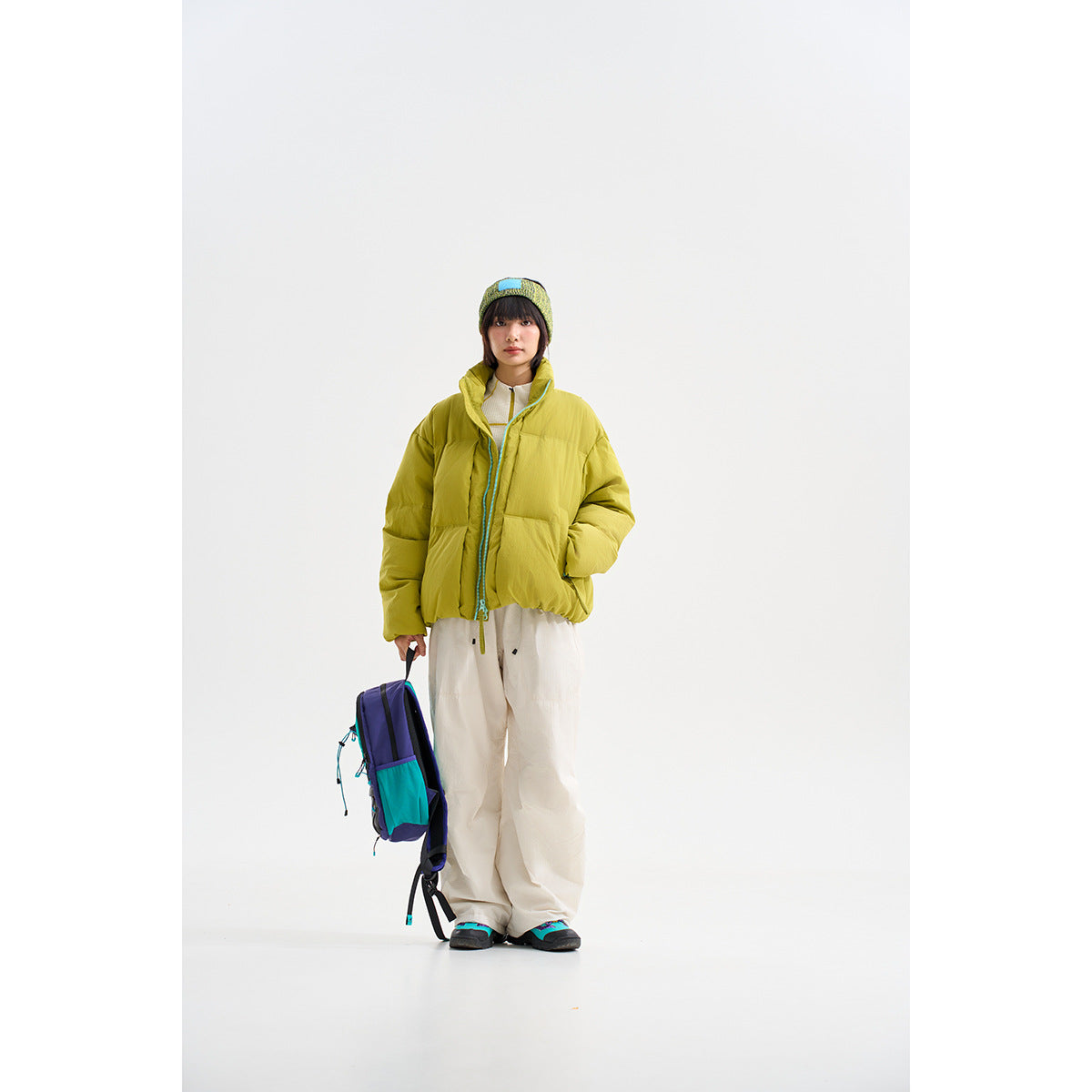Model wearing a lime-yellow puffer jacket with turquoise zipper trim, white trousers and a knit beanie.