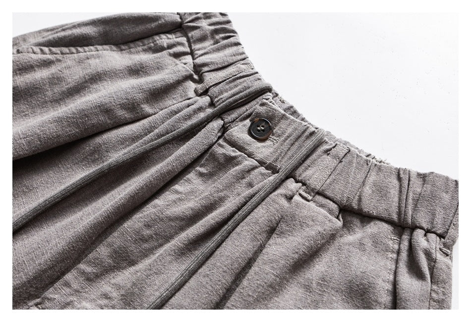 Close-up of light gray trousers showing a gathered waistband, pleats, belt loops, and a dark button.