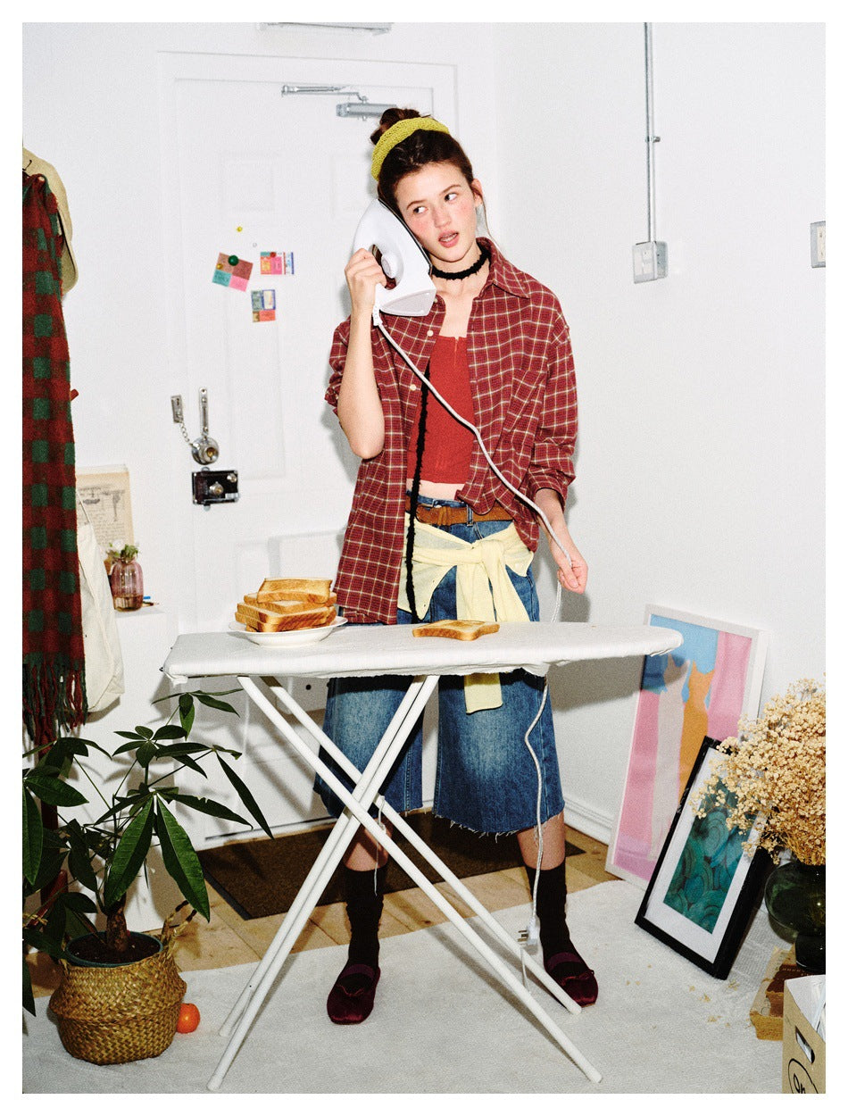 Model in an open burgundy plaid shirt over a red top and denim, holding an iron at an ironing board