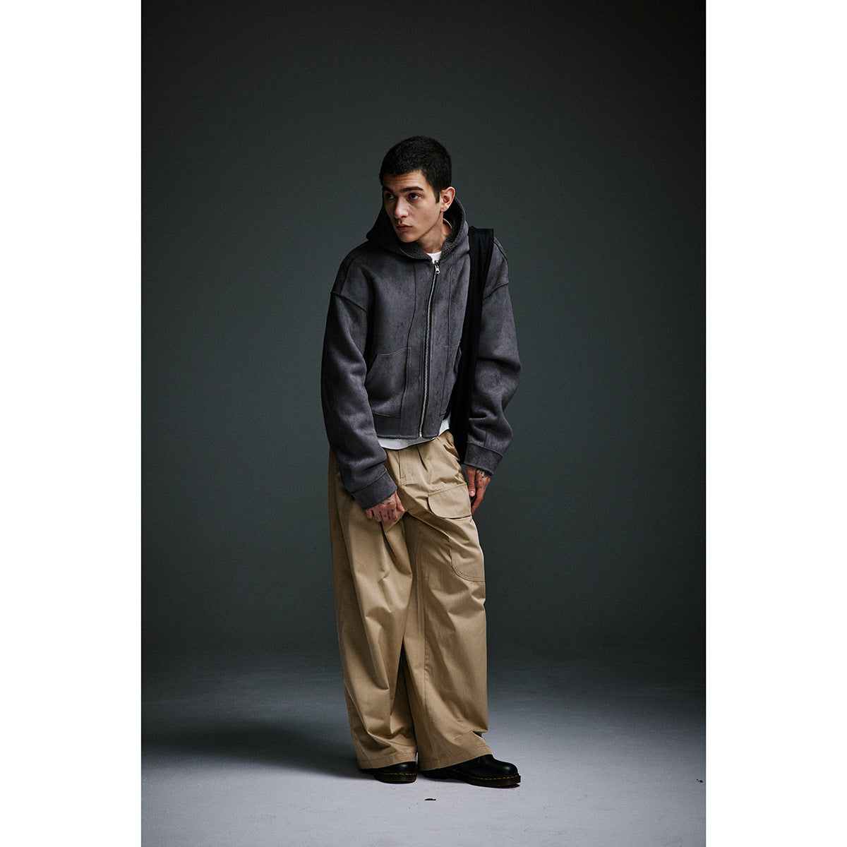 Model leaning forward in a charcoal hooded zip-up jacket and loose taupe cargo trousers.