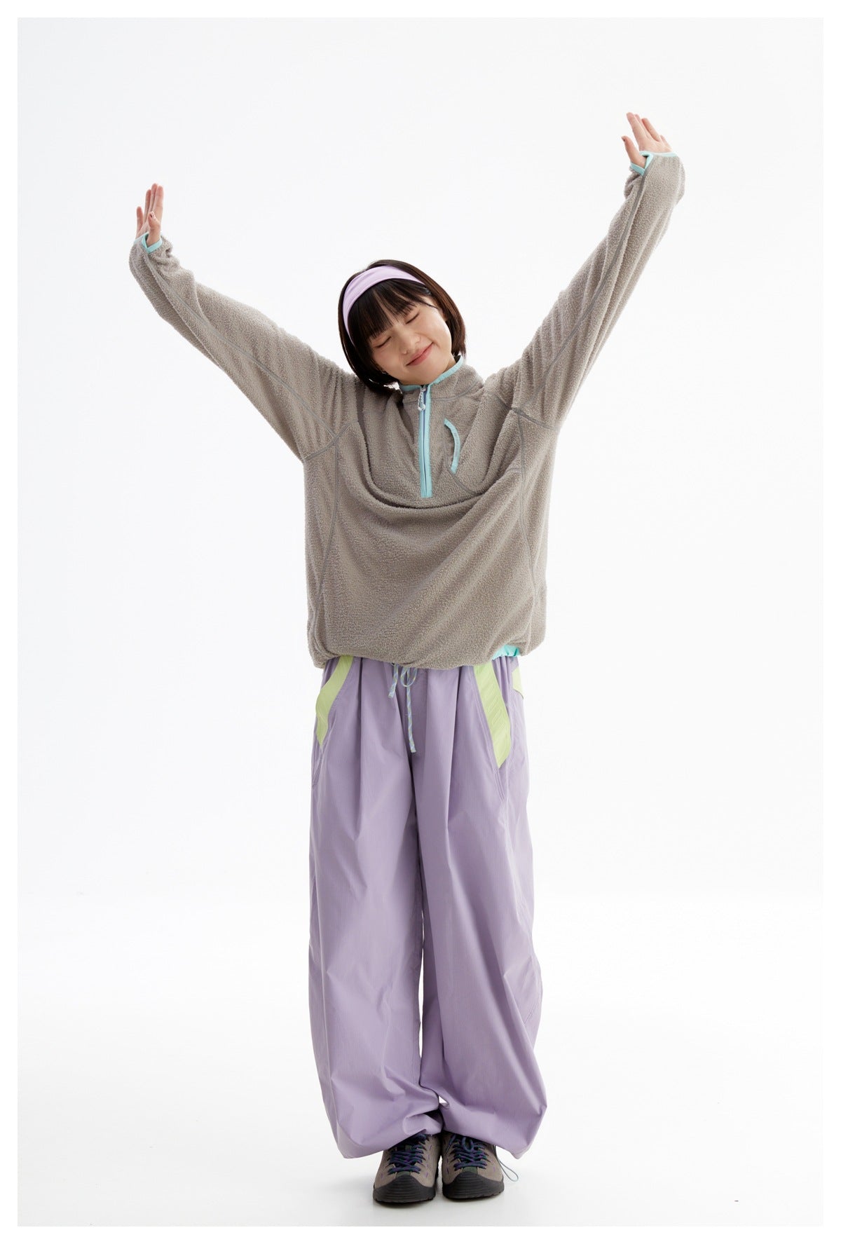 Model wearing a taupe fleece pullover with aqua trim and loose lavender pants, standing with arms raised.