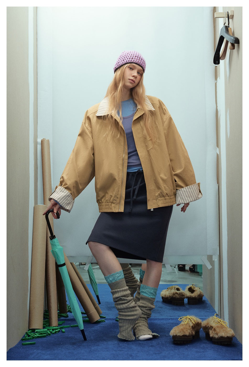 Model wearing an open tan jacket with striped collar and cuffs over blue layers and a dark skirt.