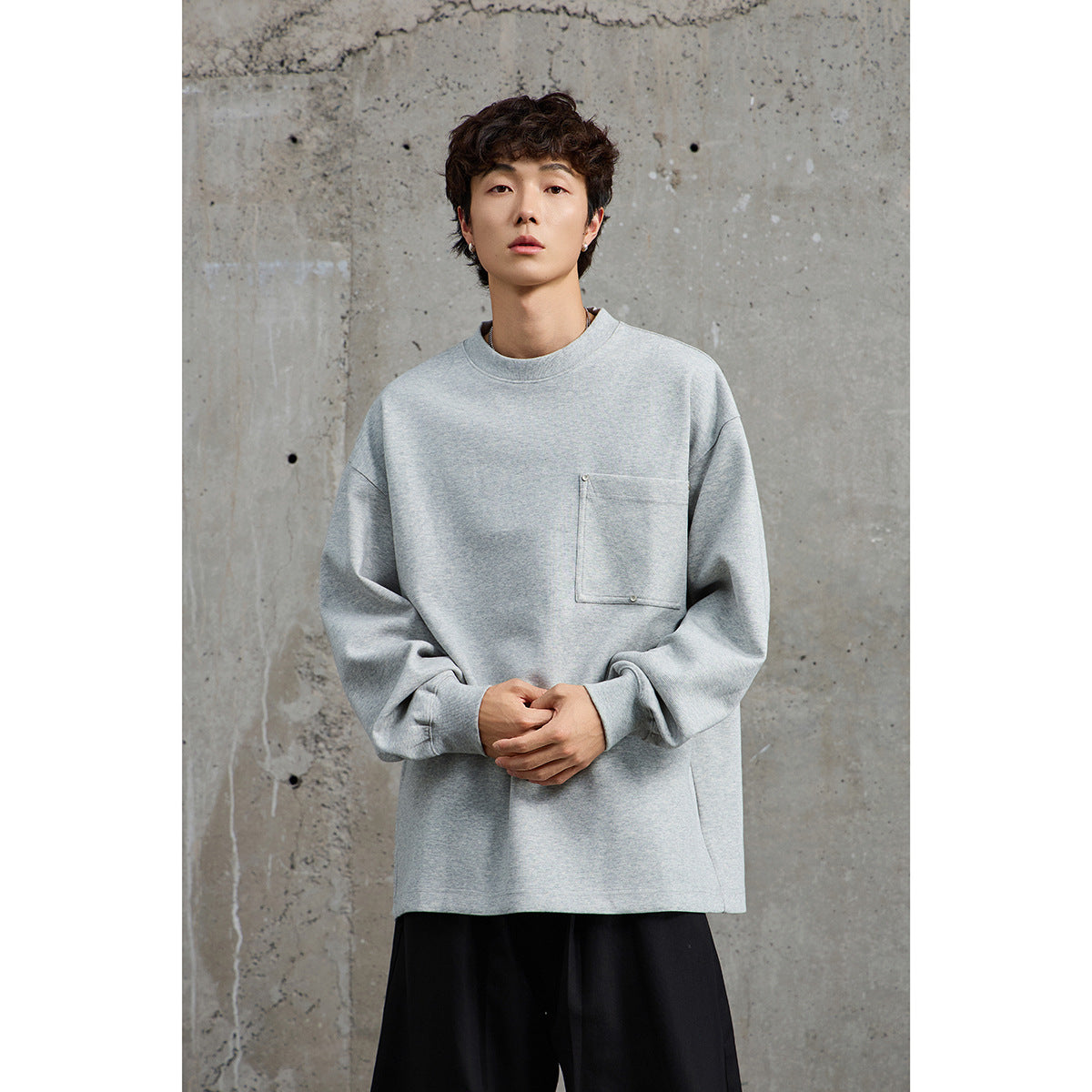 Model wearing an oversized light gray crewneck sweatshirt with a snap-detail chest pocket and black trousers.