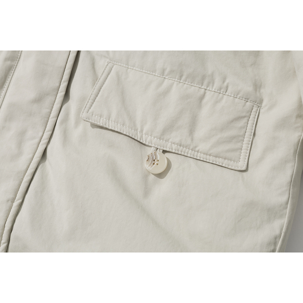 Close-up of an ivory coat’s rectangular flap pocket with a button closure and visible topstitching.