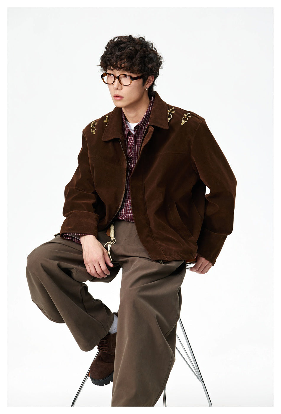 Man seated on a stool wearing a brown zip jacket over a plaid shirt with brown wide-leg pants
