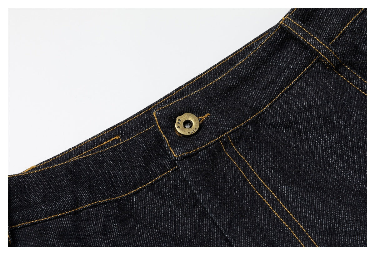 Close-up of a brass-tone button on the waistband of dark indigo jeans with ochre stitching.