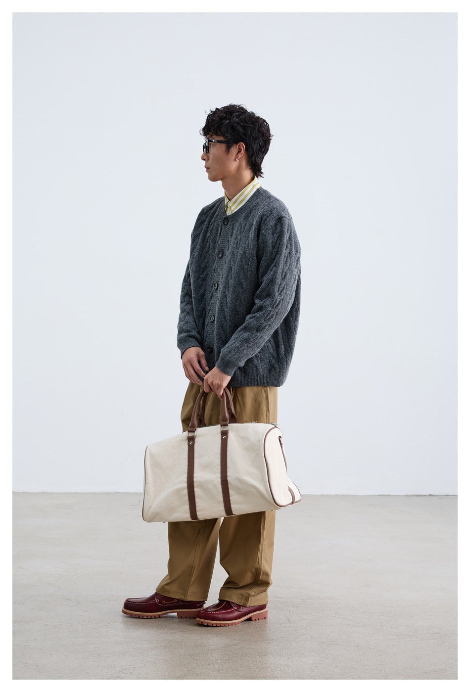 Side view of man in dark gray cable-knit cardigan over a striped shirt, holding a canvas duffel bag