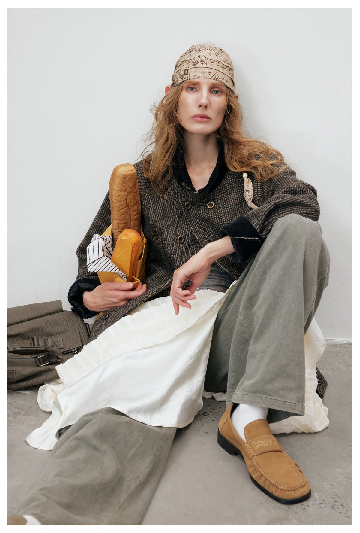 Seated model in the brown houndstooth jacket with dark collar, green pants, white skirt and a bread prop