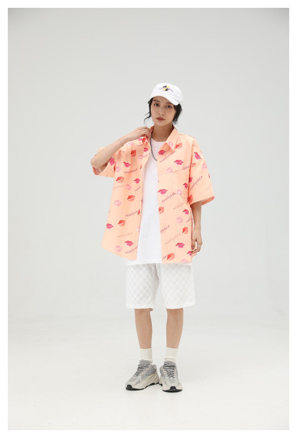 Model wearing the peach lip-print short-sleeve shirt open over a white tee with white shorts and a white cap