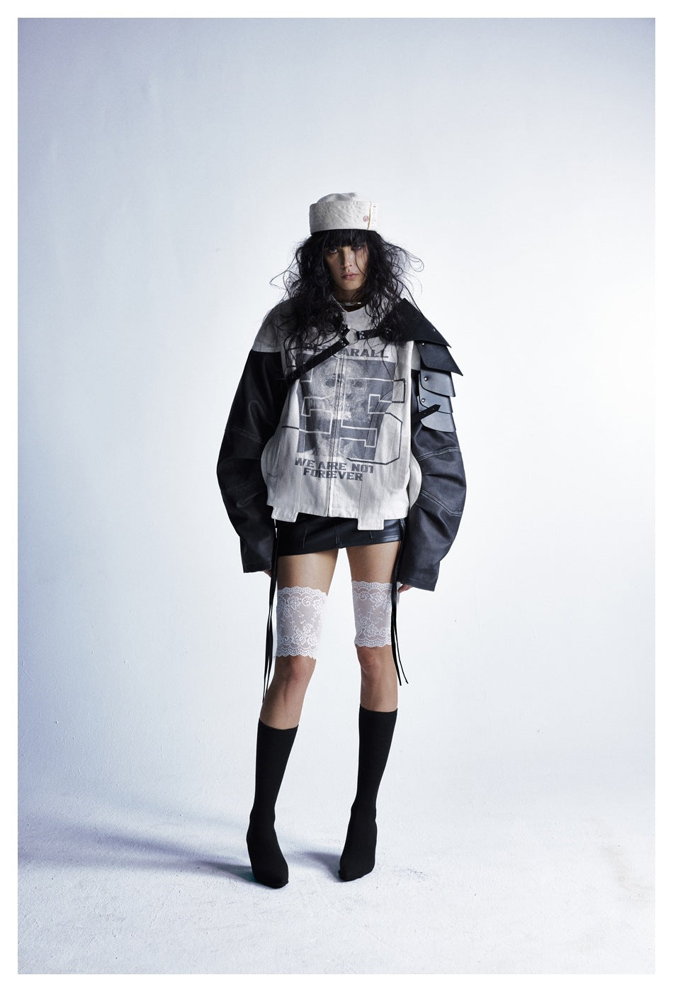 Full-length model wearing a cream-and-black graphic jacket with layered shoulder panels, a black skirt and lace thigh-highs.